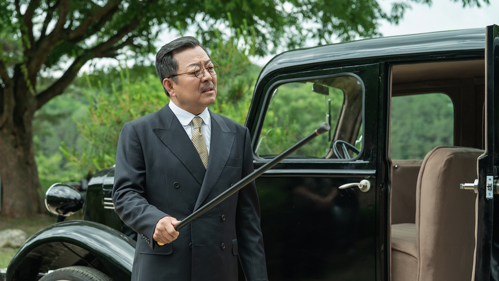 Lee Ki-young de terno risca de giz e óculos redondos, apontando uma bengala ao lado de um carro preto antigo, entre árvores