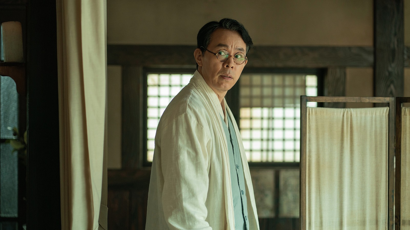 Choi Duk-moon de hanbok branco e óculos redondos, olhando por cima do ombro numa sala com janela de grade de madeira