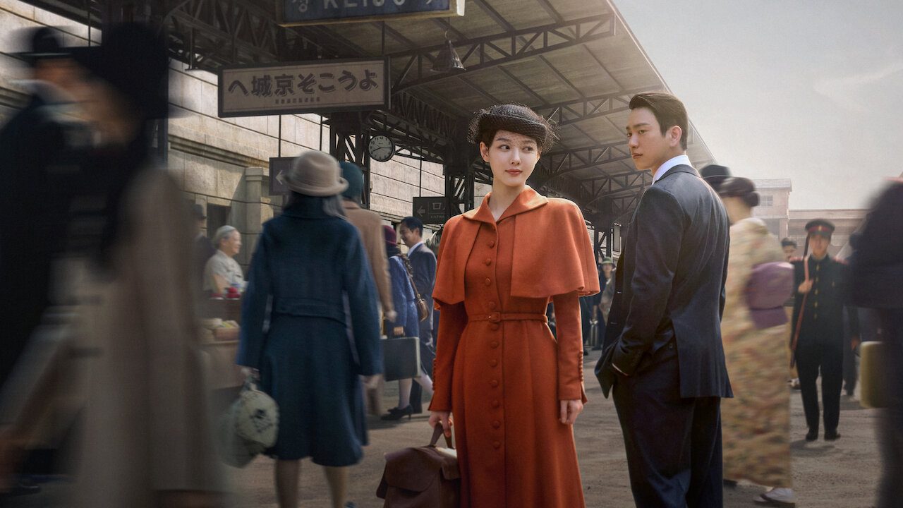 Arte oficial da série: uma moça de cabelo escuro preso sob um chapéu pequeno, vestido e capa cor de laranja, e um rapaz de terno escuro e camisa branca olhando para ela numa plataforma de estação de trem antiga, com pessoas de roupas dos anos 1930 desfocadas em volta e placa em japonês no alto
