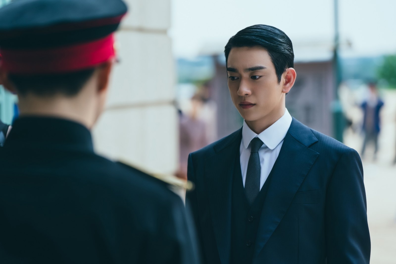 Park Jinyoung de terno azul-marinho e colete olha para baixo diante de um soldado de quepe vermelho desfocado, em cena de 100 Dias de Mentiras