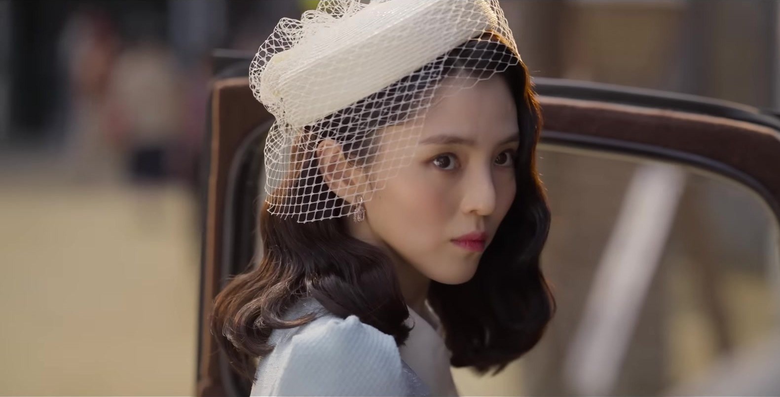 Han So-hee com chapéu branco de véu, olhando de lado, ao lado de um carro antigo, em quadro do trailer oficial de A Criatura de Gyeongseong