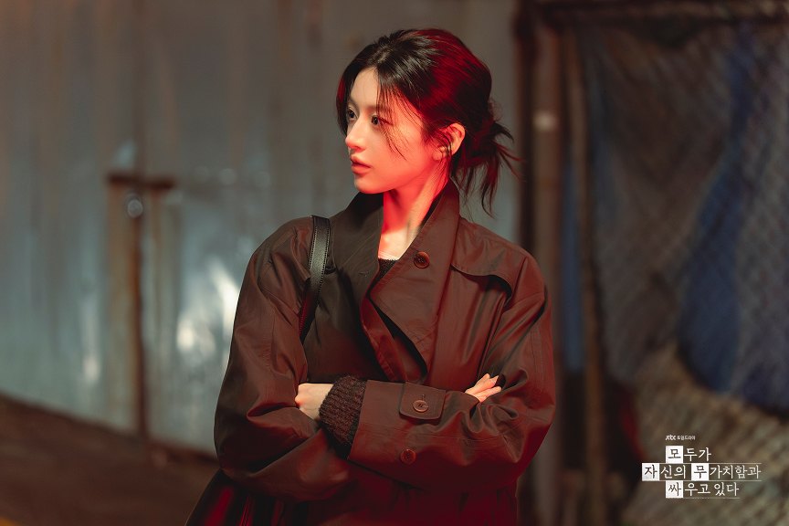 Mulher de cabelo preso e sobretudo marrom, de braços cruzados, olhando para o lado diante de uma parede de metal