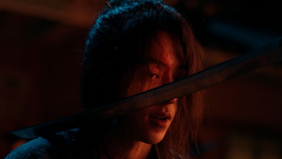 Nam Joo-hyuk, de cabelo comprido e um corte na testa, aparece na penumbra com a lâmina de uma espada atravessando a frente do rosto