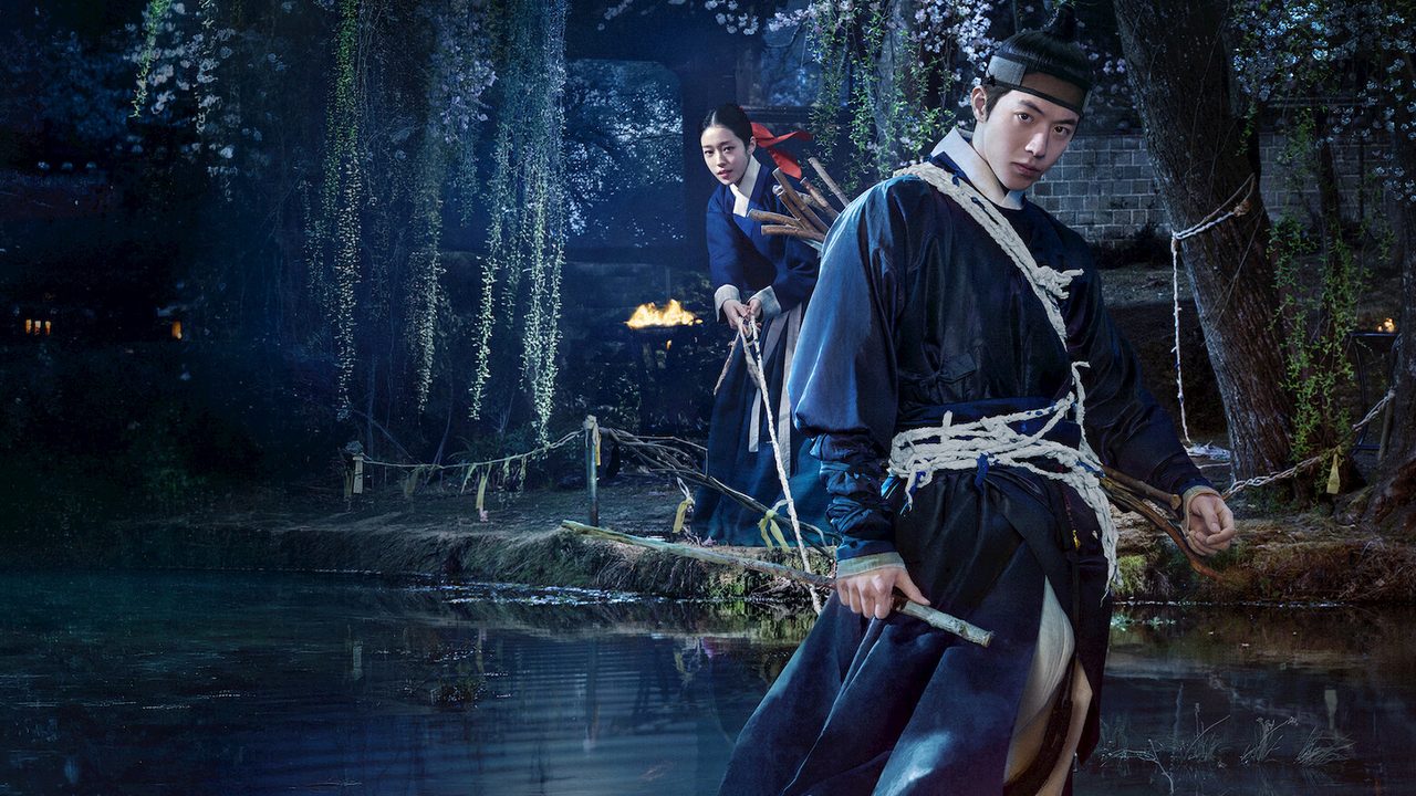 Nam Joo-hyuk, de roupa azul escura de época e chapéu, segura uma espada à beira de um lago à noite, com Roh Yoon-seo atrás dele entre árvores