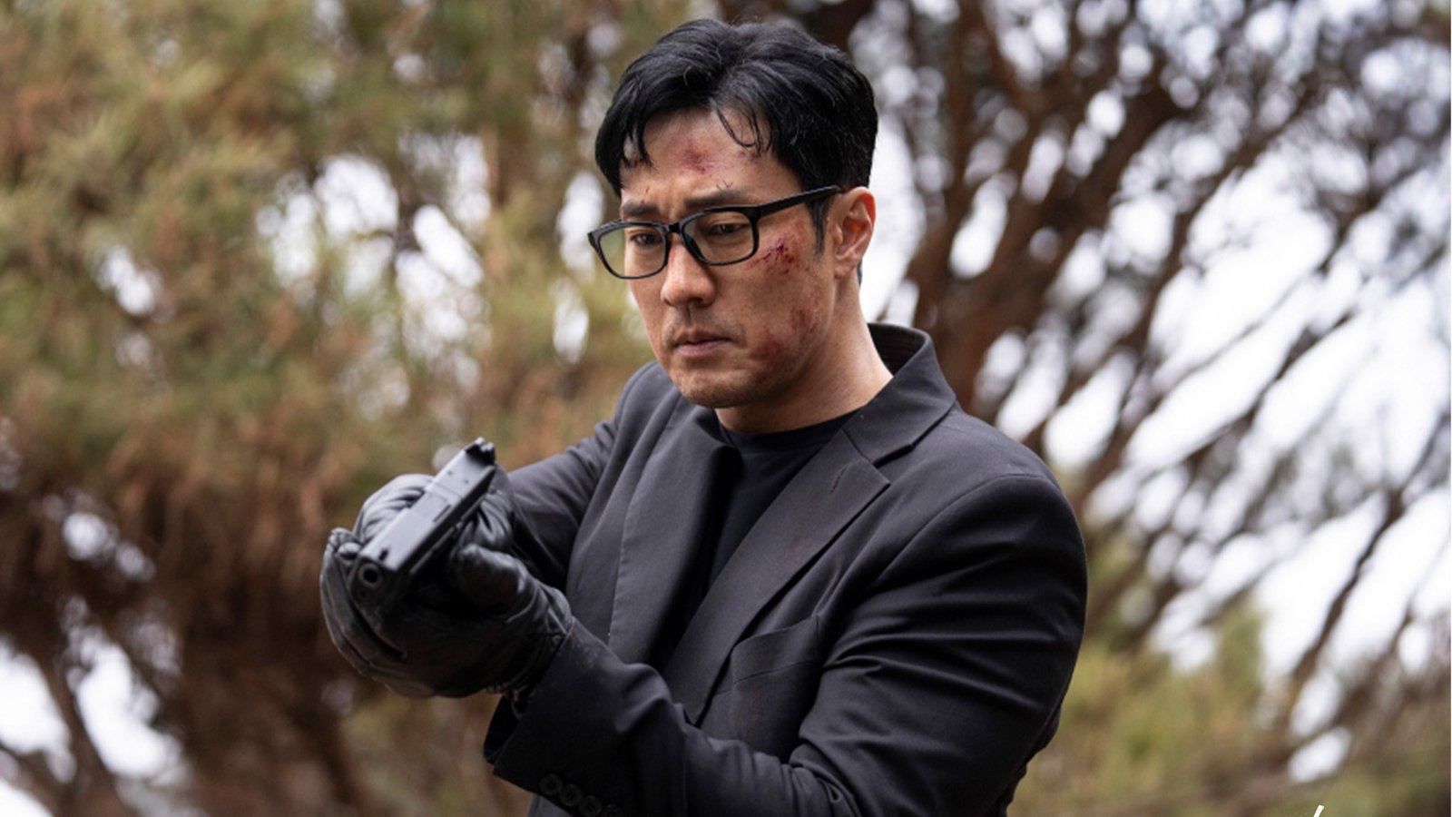 So Ji-sub como o senhor Kim, de óculos, terno preto e luvas, segurando uma pistola ao ar livre com árvores desfocadas ao fundo