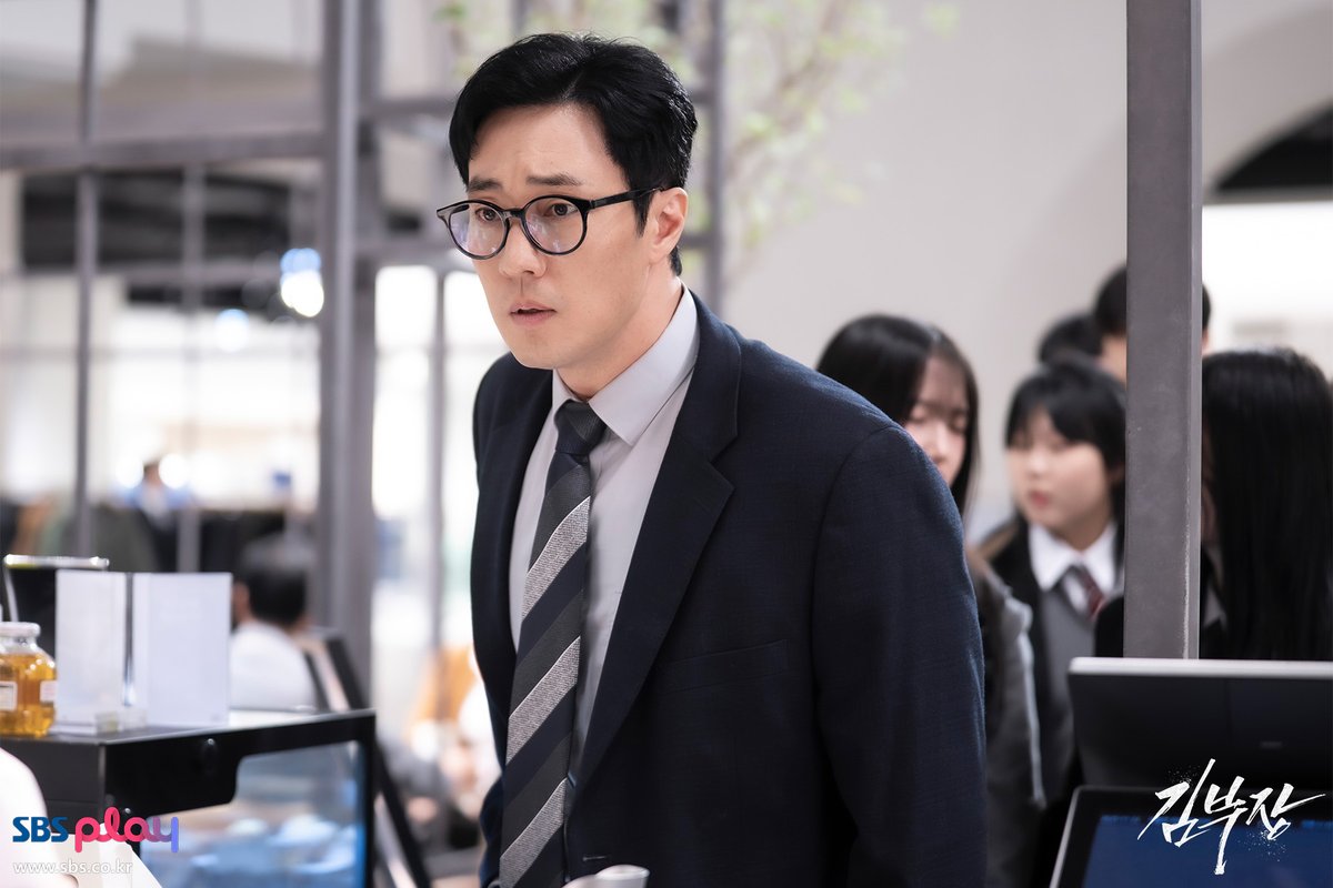 So Ji-sub de terno escuro, gravata listrada e óculos, parado no meio de uma loja movimentada, com estudantes de uniforme ao fundo
