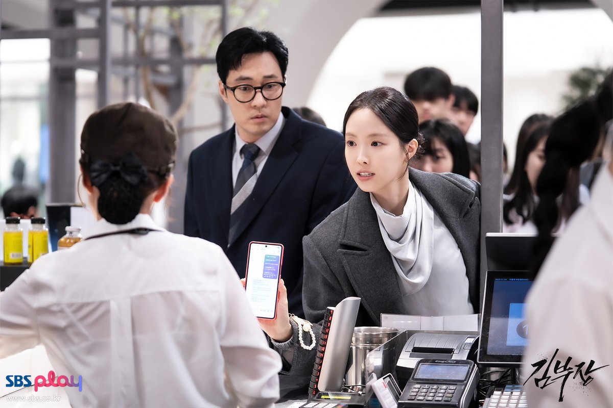 Son Na-eun de casaco cinza mostra a tela de um celular a uma atendente no balcão de uma loja, com So Ji-sub de terno e óculos logo atrás