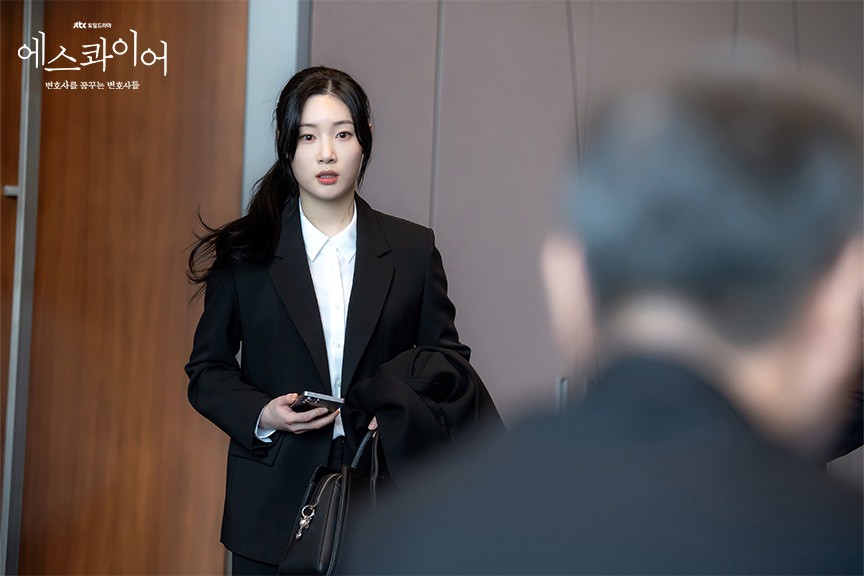 Jung Chae-yeon como Hyo-min, de terno preto e camisa branca, segura o celular e um casaco num corredor de paredes de madeira