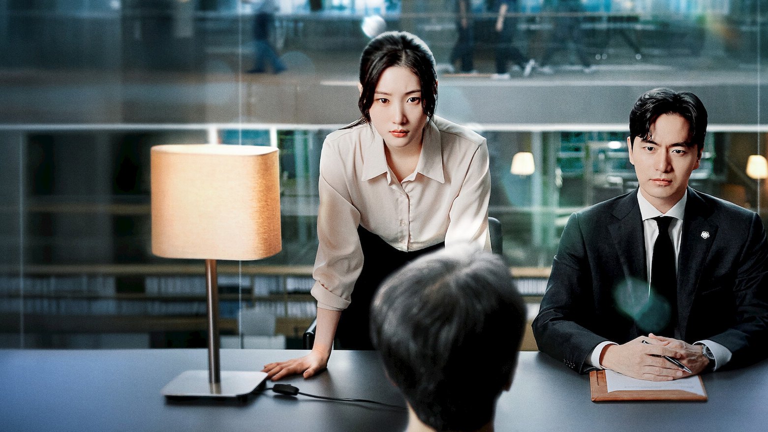 Jung Chae-yeon, de camisa clara, se inclina sobre uma mesa de reunião ao lado de Lee Jin-uk, de terno escuro, diante de uma pessoa de costas