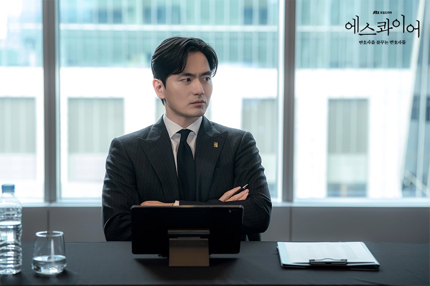 Lee Jin-uk como Seok-hoon, de terno risca de giz e gravata preta, está sentado de braços cruzados atrás de uma mesa com tablet, pasta e copo d'água