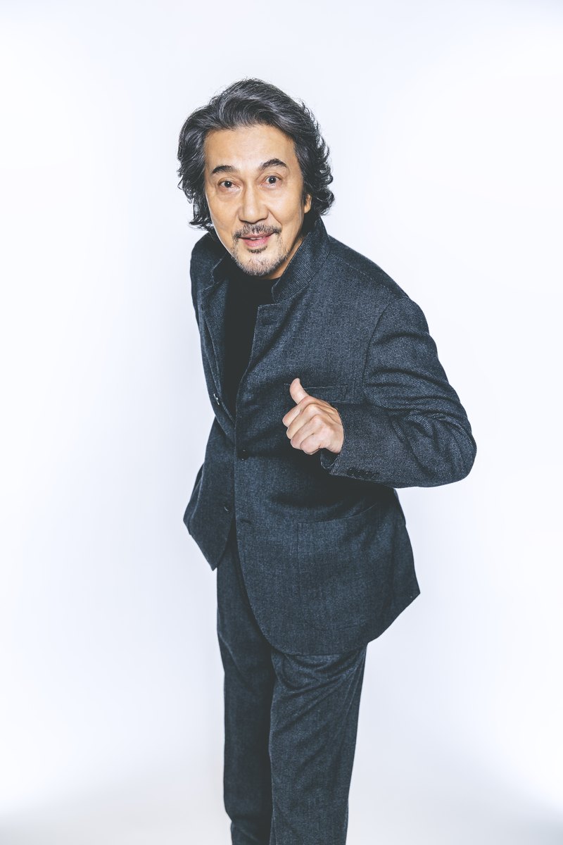 Koji Yakusho de terno escuro, apontando para si mesmo, em fundo branco