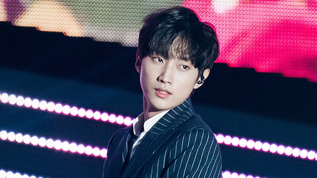 Jin Young de terno risca de giz escuro, olhando para a frente, diante de painel de luzes no palco