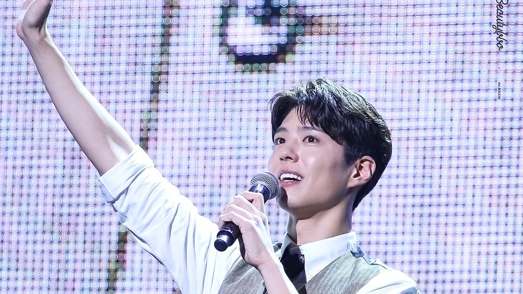Park Bo Gum de colete cinza e camisa branca acena com o braço levantado e segura um microfone no palco