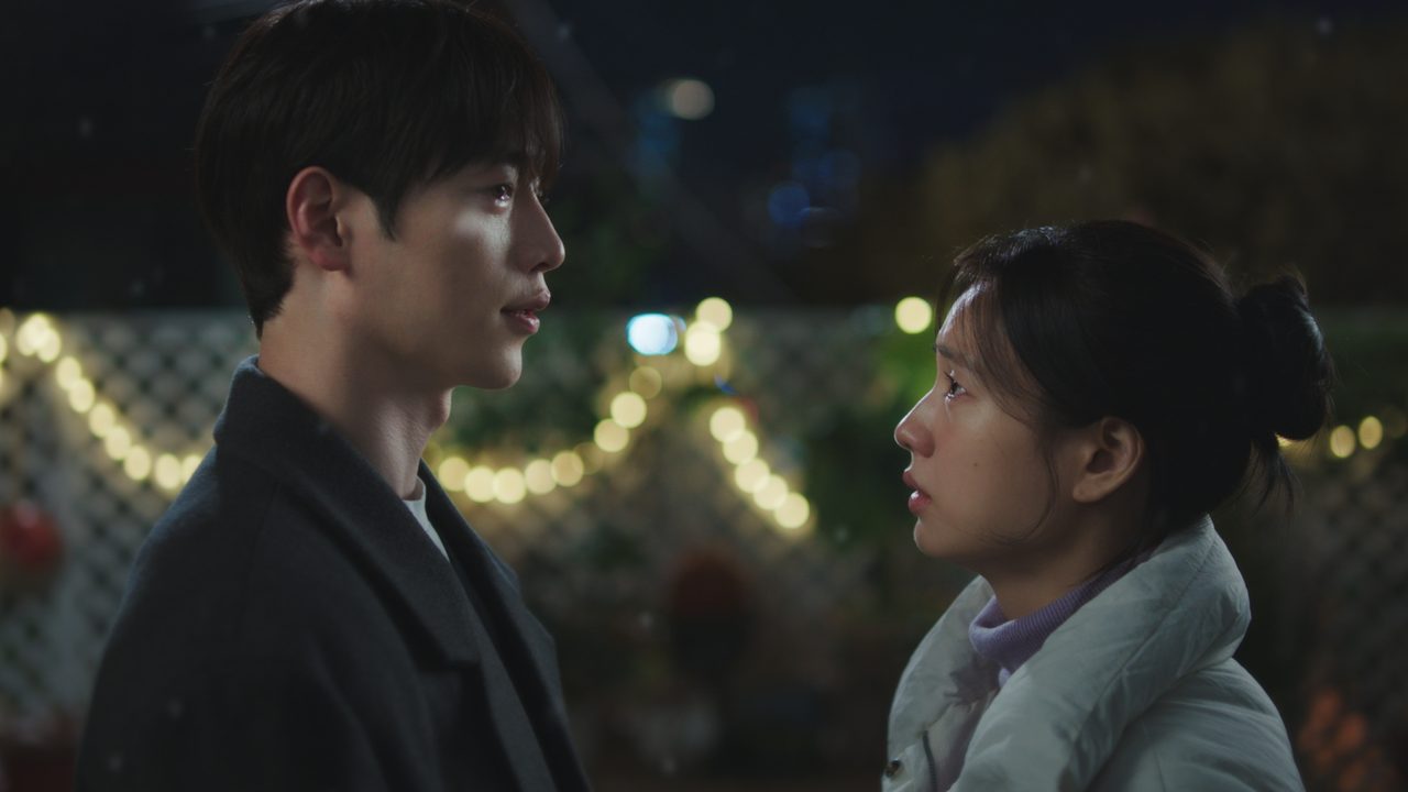 Seo Kang-joon, de casaco escuro, e Ahn Eun-jin, de jaqueta branca, frente a frente à noite num quintal com cordão de luzes ao fundo