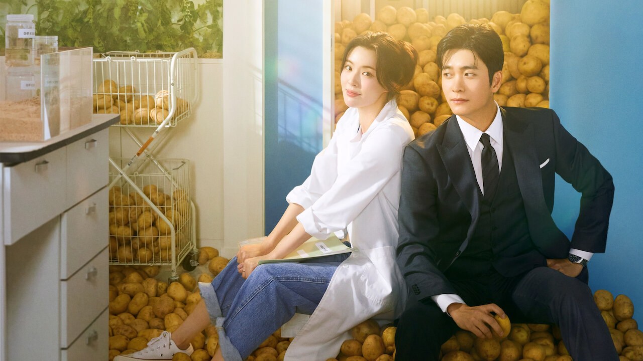 Arte de divulgação de Amor e Batatas na Netflix: mulher de jaleco branco e jeans sentada ao lado de um homem de terno escuro e gravata, com prateleiras de batatas ao fundo e uma bancada de laboratório à esquerda