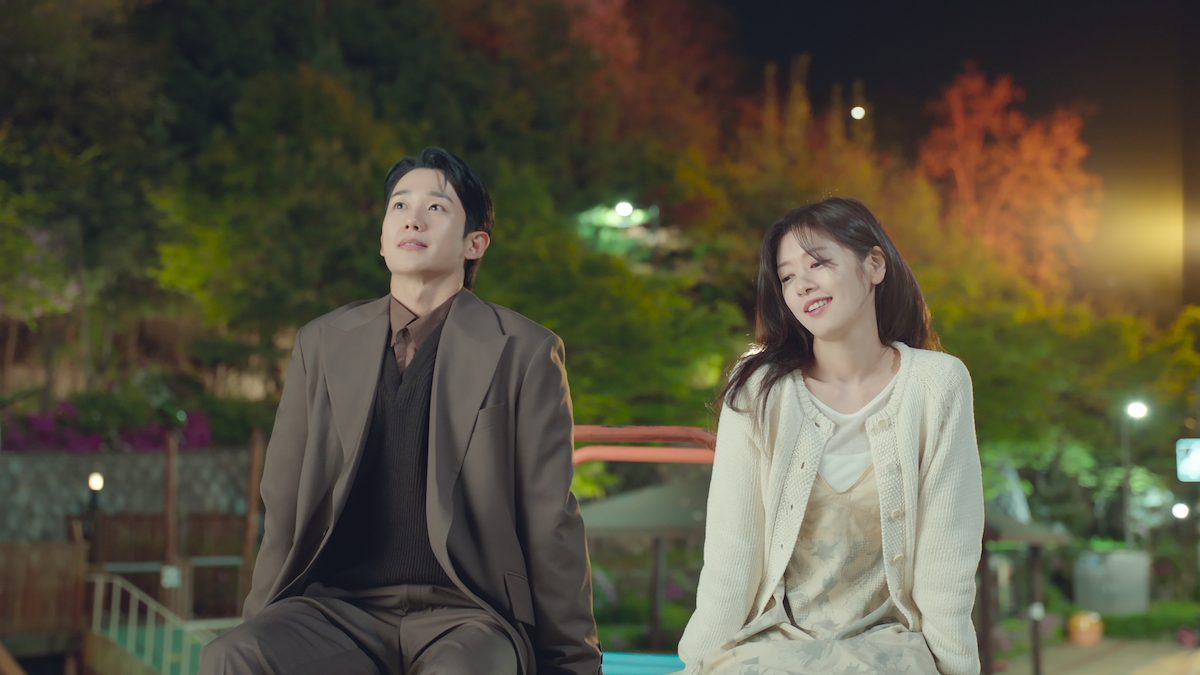 Jung Hae-in de terno marrom e Jung So-min de vestido florido e cardigã creme sentados num banco à noite, sob árvores iluminadas