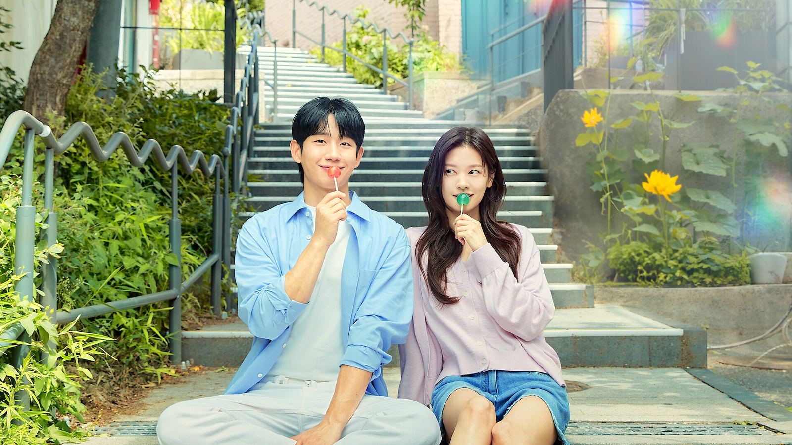 Jung Hae-in de camisa azul e Jung So-min de cardigã lilás sentados no pé de uma escadaria ao ar livre, cada um com um pirulito, entre plantas e flores amarelas