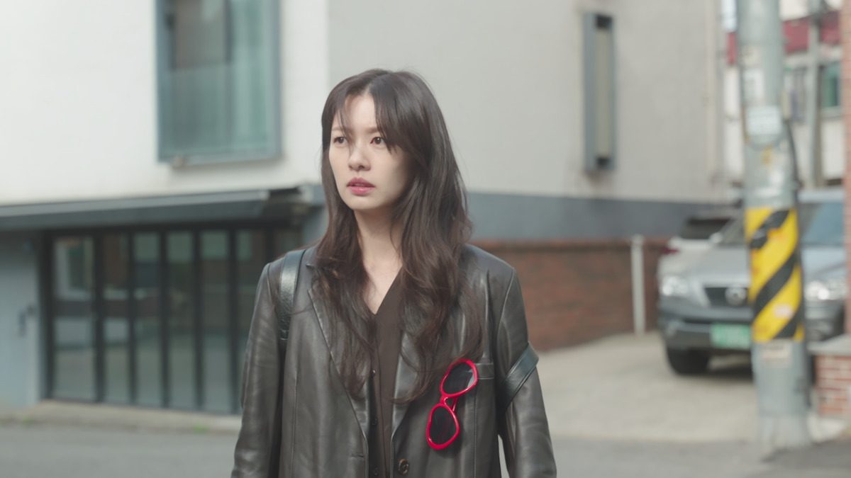 Jung So-min como Bae Seok-ryu, de jaqueta de couro preta com óculos vermelhos em forma de coração presos no bolso, parada numa rua diante de um prédio cinza