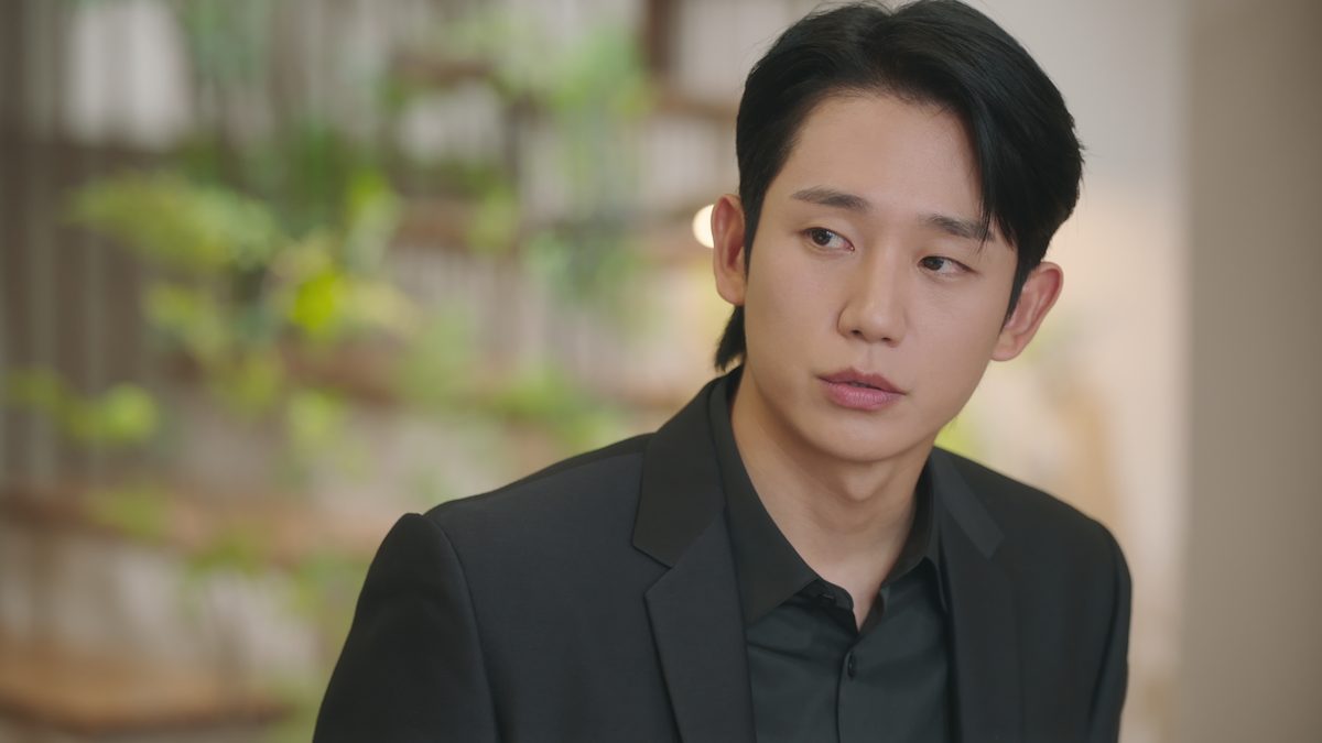 Jung Hae-in como Choi Seung-hyo, de paletó e camisa pretos, olhando de lado com expressão séria diante de folhagem desfocada