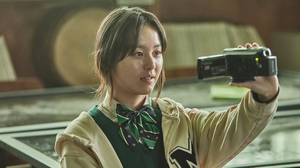 Park Ji-hu como Nam On-jo, de uniforme com gravata-borboleta verde e jaqueta clara, segurando uma filmadora num laboratório escolar