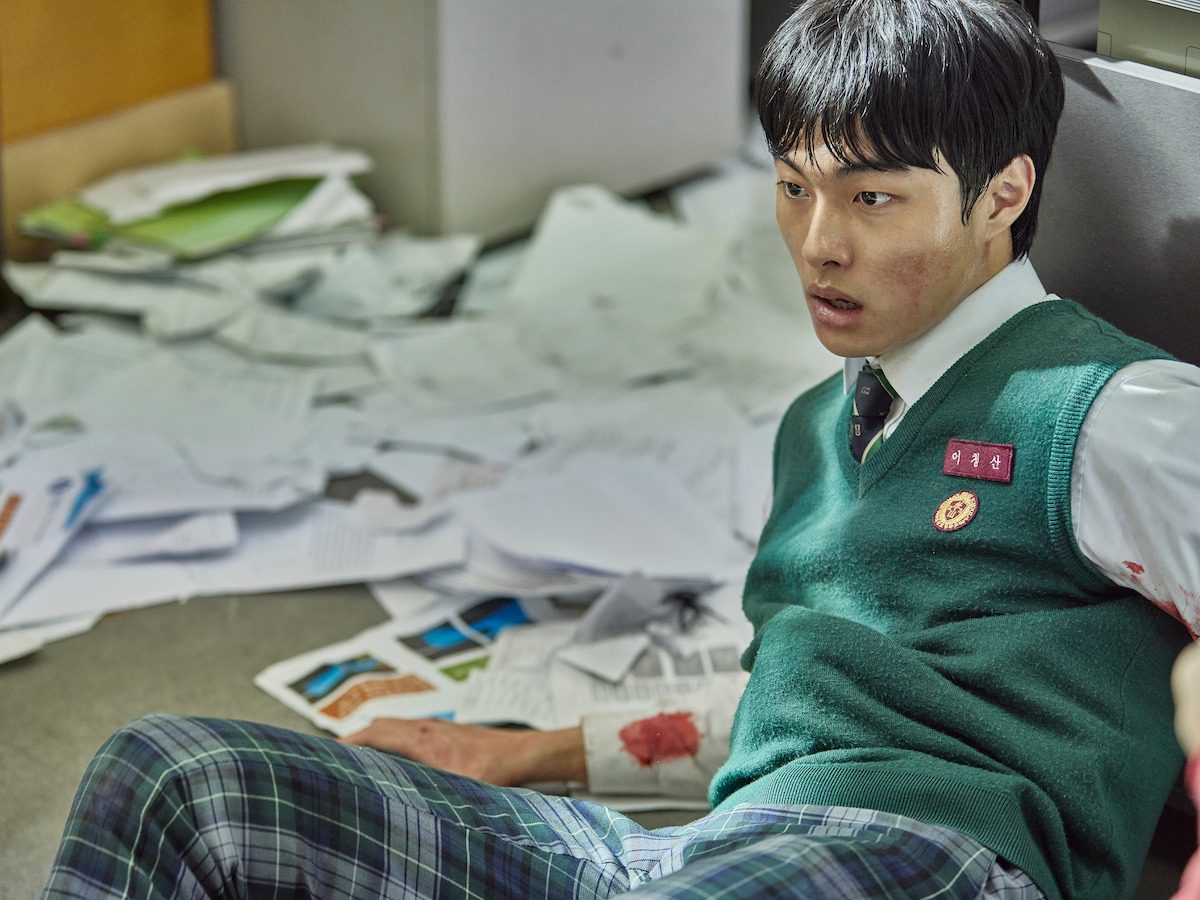 Yoon Chan-young como Lee Cheong-san, de colete verde do uniforme e crachá com o nome, sentado no chão entre papéis espalhados