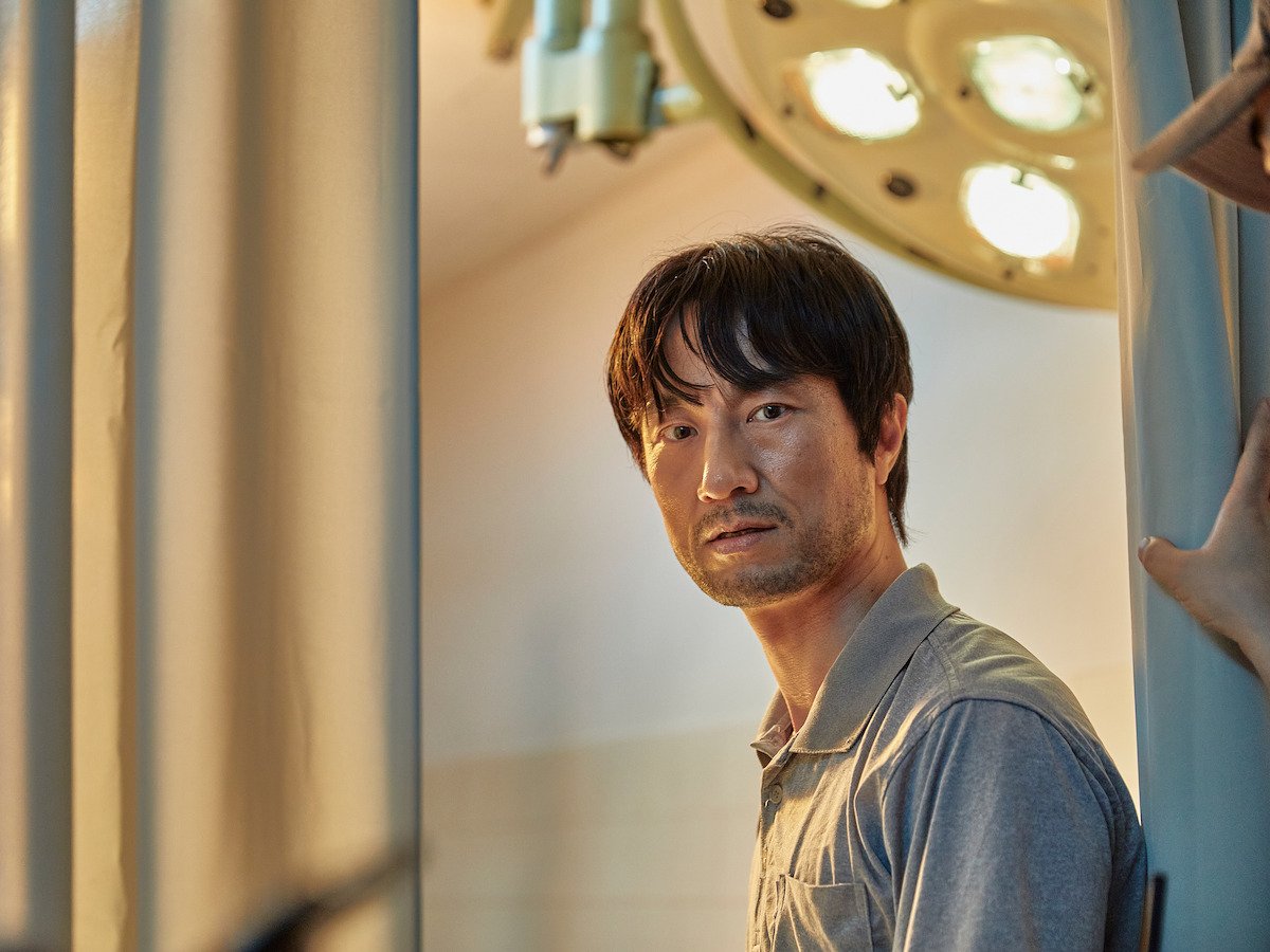 Kim Byong-chul como o professor Lee Byeong-chan, de camisa cinza, olhando para o lado num laboratório com luminária cirúrgica e persianas