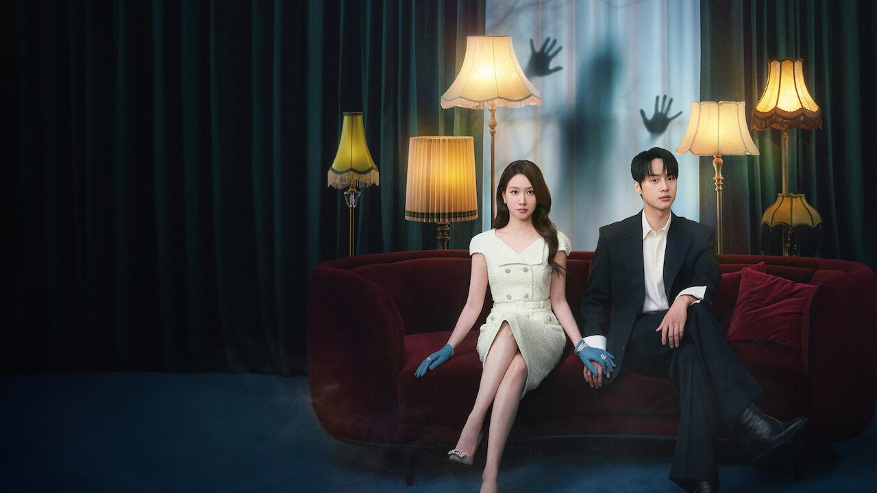 Park Eun-bin de vestido claro e luvas azuis e Yang Se-jong de terno preto de mãos dadas num sofá de veludo vermelho, entre abajures, com sombras de mãos na cortina ao fundo