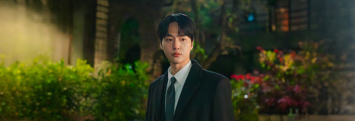 Yang Se-jong como Ma Gang-uk, de terno preto, camisa branca e gravata escura, parado num jardim à noite entre arbustos e flores vermelhas