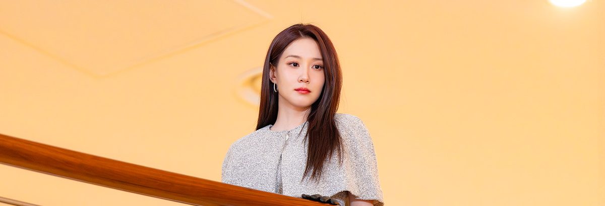 Park Eun-bin como Cheon Yeo-ri, de blusa prateada de tweed e argola na orelha, debruçada num corrimão de madeira sob teto amarelo, com a mão de luva escura apoiada