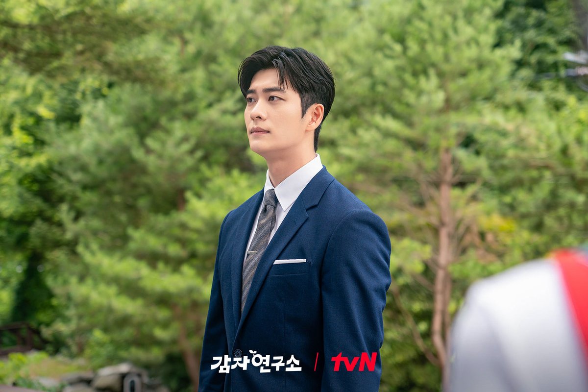 Kang Tae-oh como So Baek-ho, de terno azul-marinho e gravata cinza, olha para o lado num jardim cercado de pinheiros