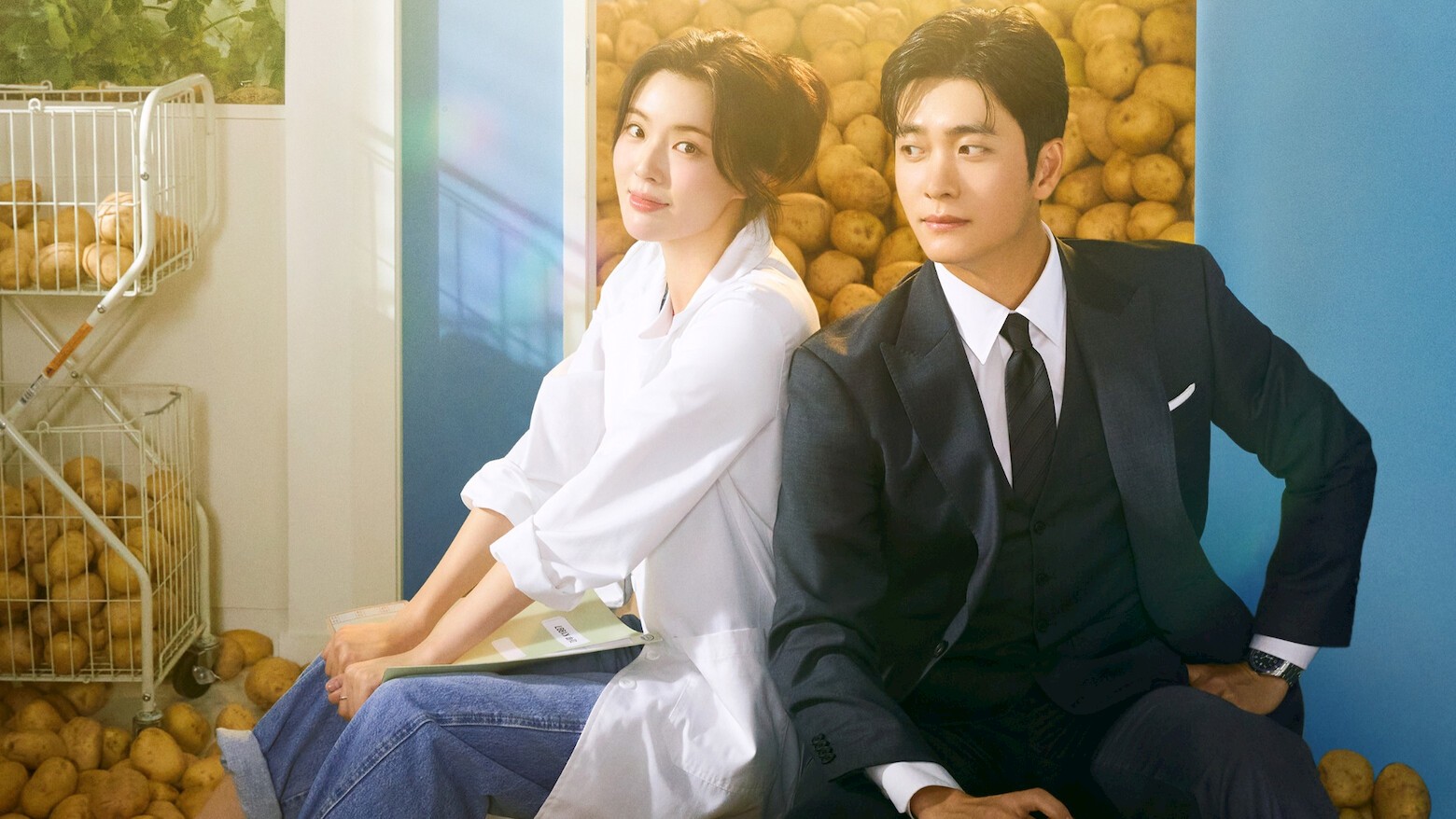 Lee Sun-bin de jaleco branco e jeans e Kang Tae-oh de terno escuro sentados de costas um para o outro diante de uma parede de batatas