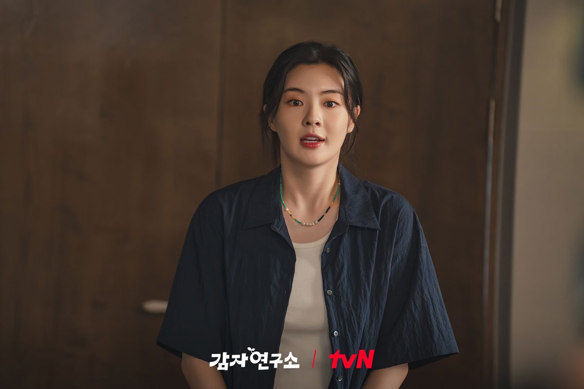 Lee Sun-bin como Kim Mi-kyung, de camisa azul-marinho aberta sobre blusa clara e colar de contas, olha surpresa diante de uma porta de madeira