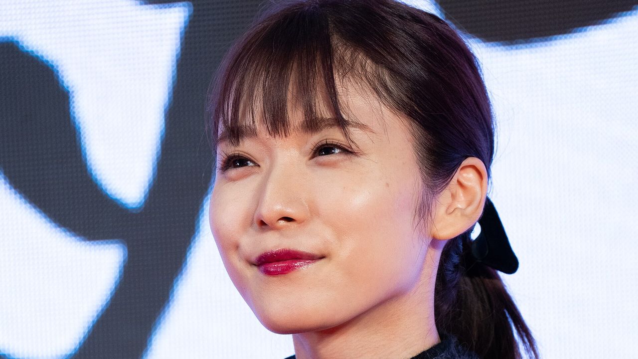 Mayu Matsuoka sorri de franja e cabelo preso com laço preto, diante de um painel claro com letras pretas