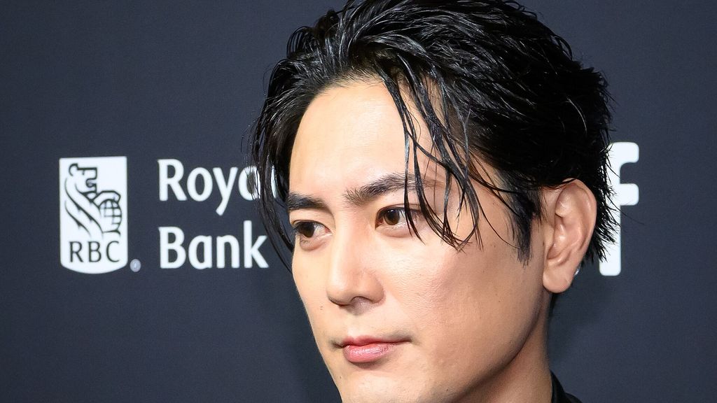 Shotaro Mamiya de cabelo escuro penteado para trás, sério, diante do painel de patrocinadores do festival