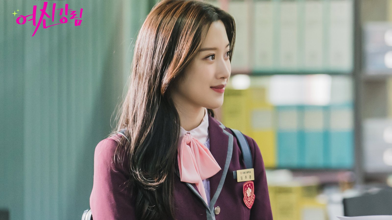 Moon Ga-young como Joo-gyeong, de uniforme vinho com laço rosa e crachá, sorri de perfil numa sala com estantes de pastas coloridas