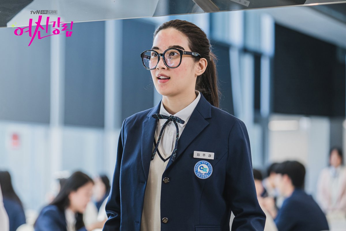Moon Ga-young como Joo-gyeong, de óculos redondos de aro grosso, rabo de cavalo e uniforme azul-marinho com laço, num refeitório escolar