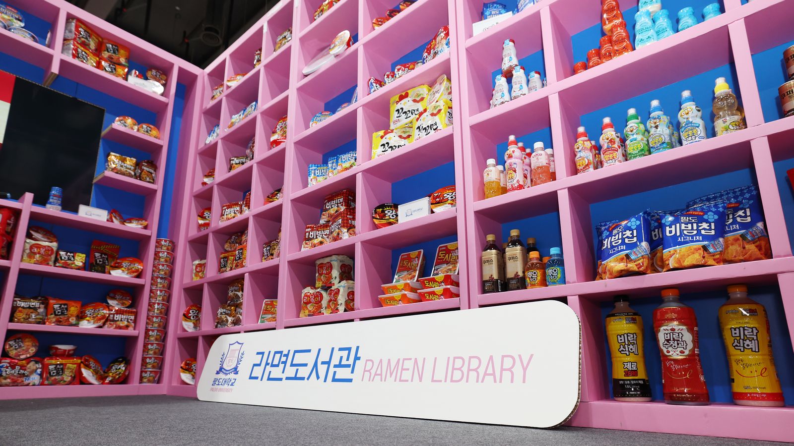 Estantes rosa com pacotes de ramyeon, salgadinhos e garrafas de bebida num estande de feira, com a placa Ramen Library no chão