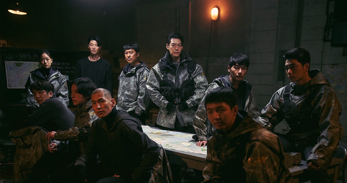 Dez pessoas com jaquetas camufladas e roupas escuras reunidas em volta de uma mesa coberta de mapas, numa sala de concreto com luminárias acesas; Kim Woo-bin de pé ao centro