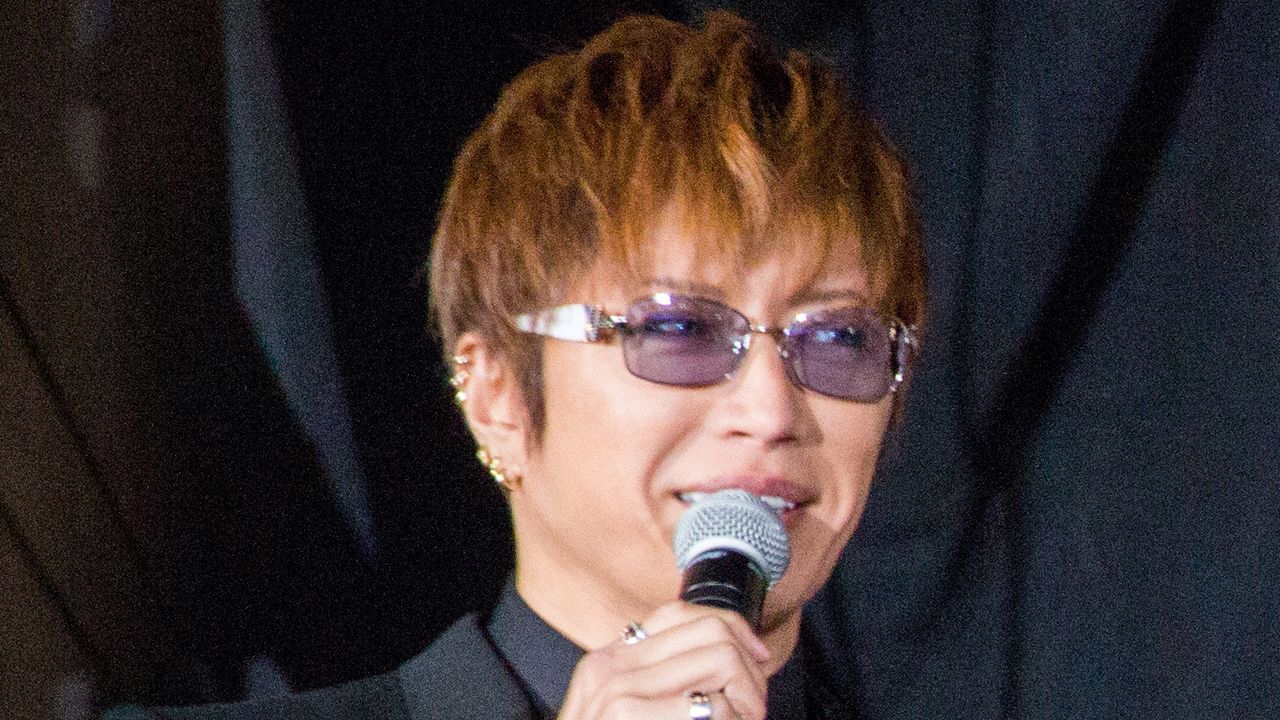 GACKT, de óculos de lentes lilás e paletó preto, fala ao microfone diante de uma cortina escura