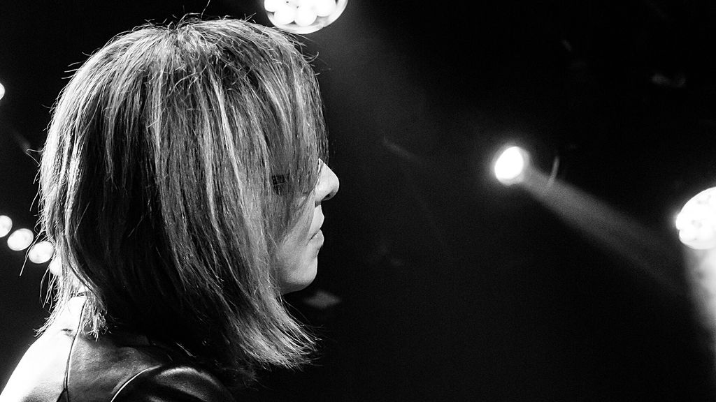 YOSHIKI, de perfil e cabelo claro na altura do queixo, em foto em preto e branco com refletores ao fundo