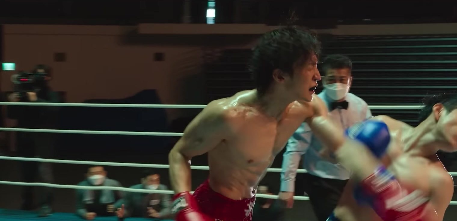 Boxeador sem camisa, de calção vermelho, acerta um soco no ringue com o árbitro de máscara ao fundo, em quadro do trailer oficial de Bloodhounds