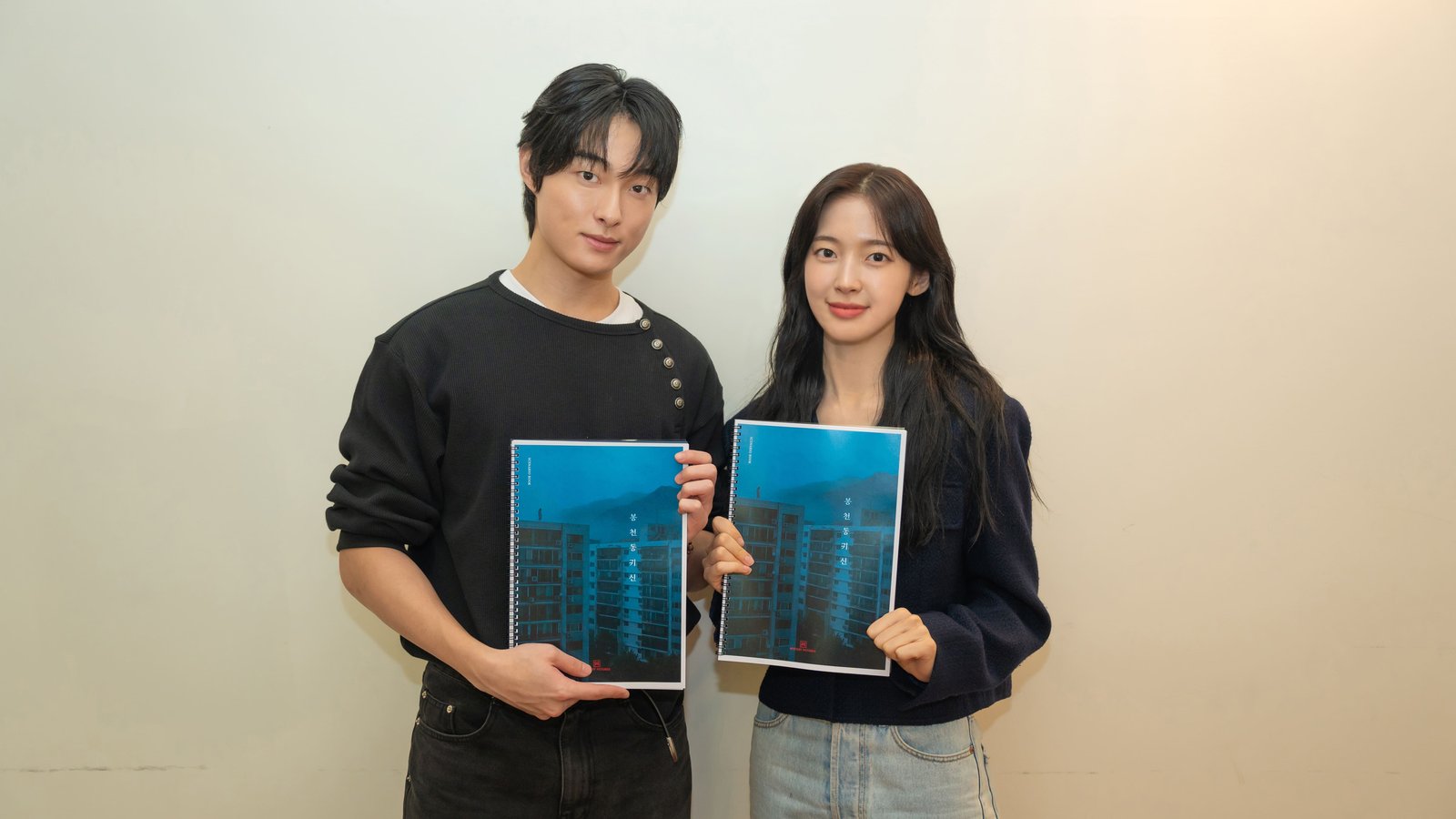 Yoon Chan Young, de suéter preto, e Arin, de cardigã azul e jeans claro, lado a lado diante de parede bege, cada um segurando o roteiro de capa azul do filme com um prédio e o título em coreano