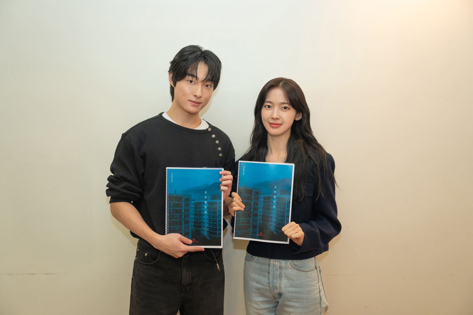 Yoon Chan Young e Arin lado a lado segurando o roteiro azul de Bongcheon-dong Ghost, com um prédio na capa