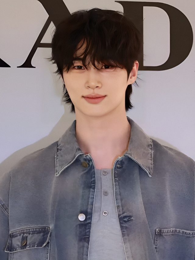 Byeon Woo-seok de jaqueta jeans e camiseta cinza sorrindo diante de painel claro