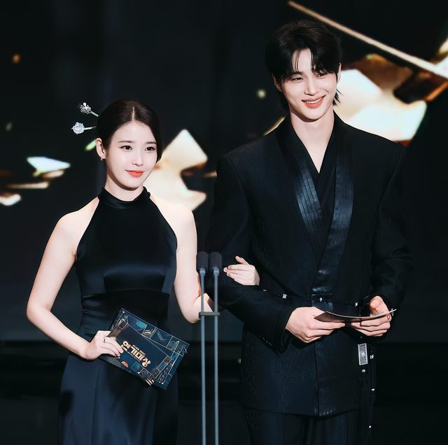IU de vestido preto e Byeon Woo-seok de terno preto no palco de uma premiação, com envelope e cartão nas mãos