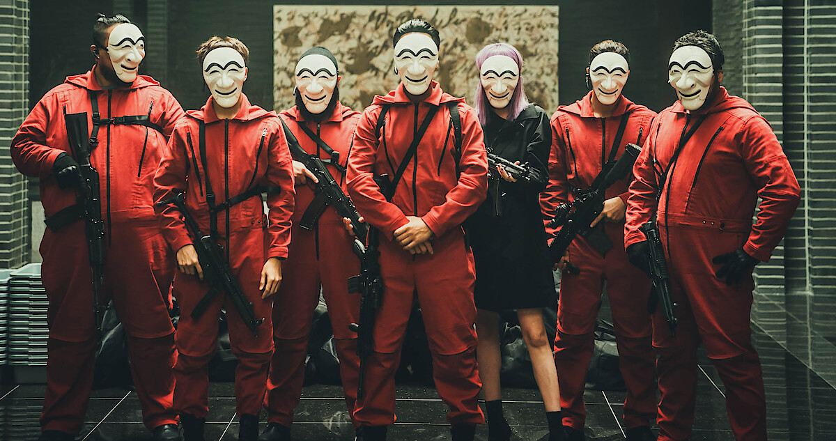 Sete pessoas armadas com máscaras brancas de estilo Hahoe, seis de macacão vermelho e uma de roupa preta e cabelo lilás, enfileiradas diante de uma parede