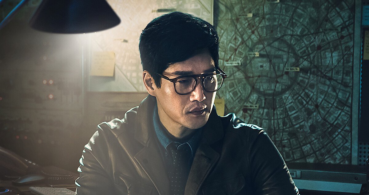 Yoo Ji-tae como o Professor, de óculos, jaqueta escura e gravata, sob uma luminária, diante de uma parede coberta de mapas e papéis