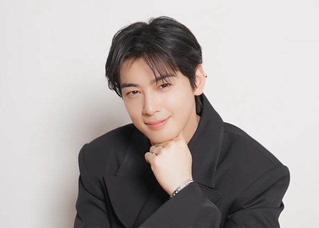 Cha Eun-woo sorrindo com o queixo apoiado na mão, de blazer preto, em fundo branco