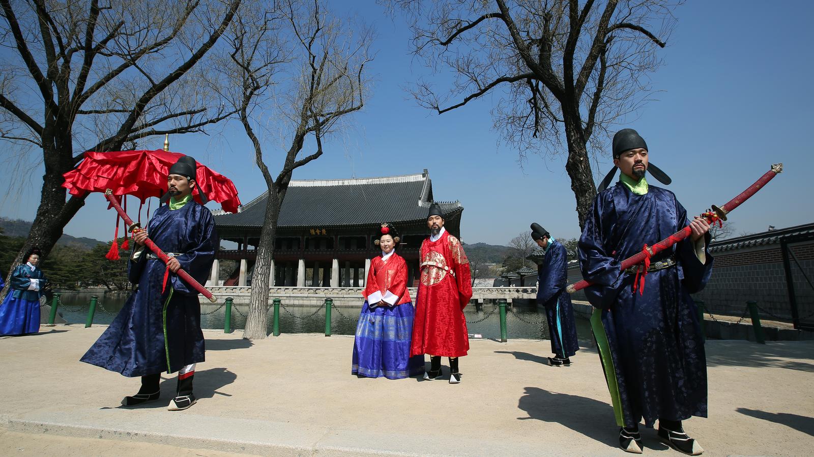 Participantes em trajes tradicionais coreanos, com chapéus pretos, guarda-sol vermelho e espadas de bainha vermelha, diante de um pavilhão do palácio