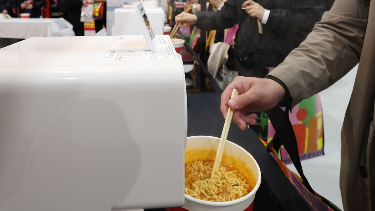 Mão de visitante segura hashis sobre um copo de ramyeon ao lado de uma máquina branca, com outras pessoas ao fundo